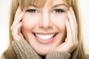 Smiling blonde person with hands on cheeks, wearing a beige sweater, close-up portrait