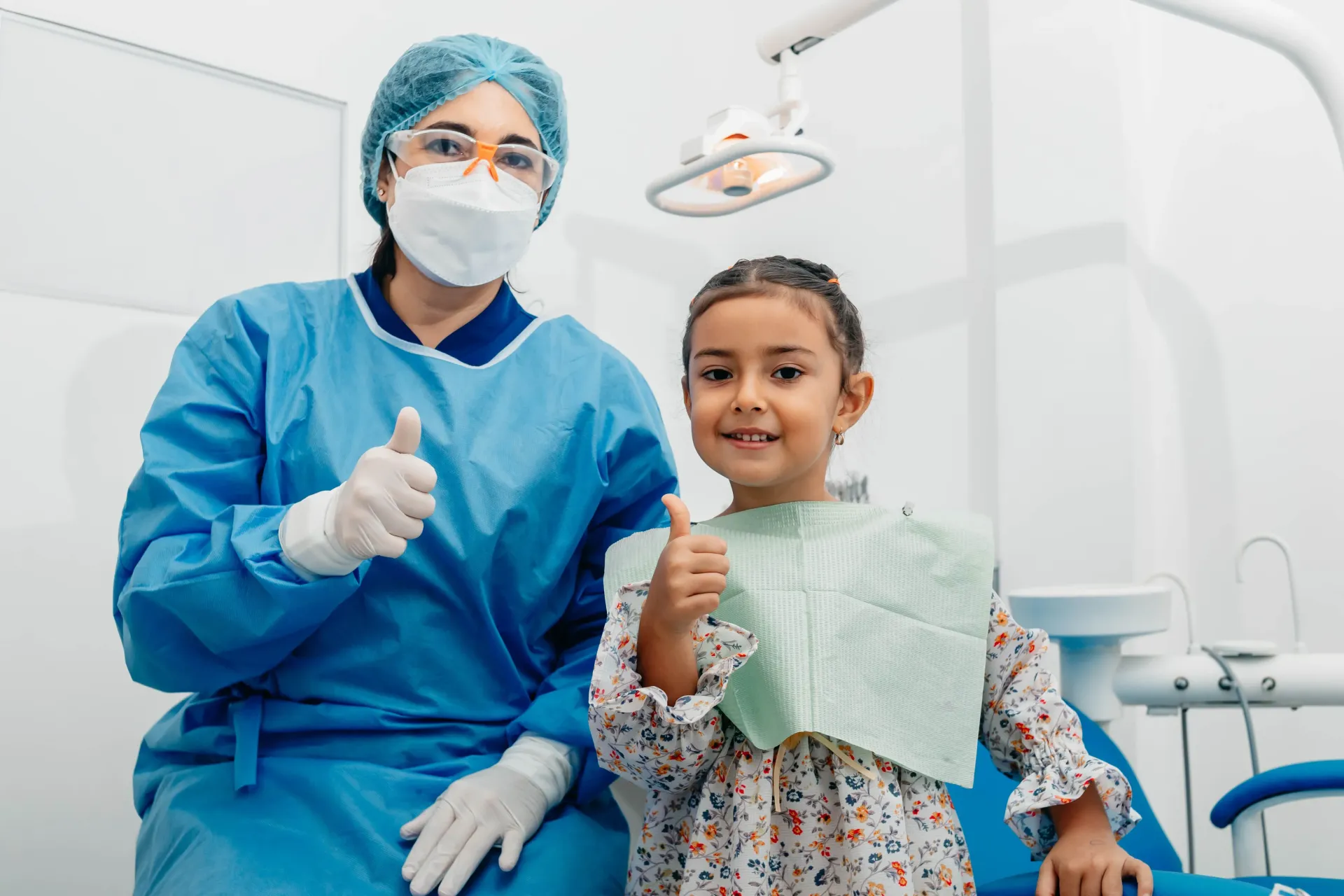 Dentist and child in a clinic, both giving thumbs up, with a dental chair and equipment in the background.