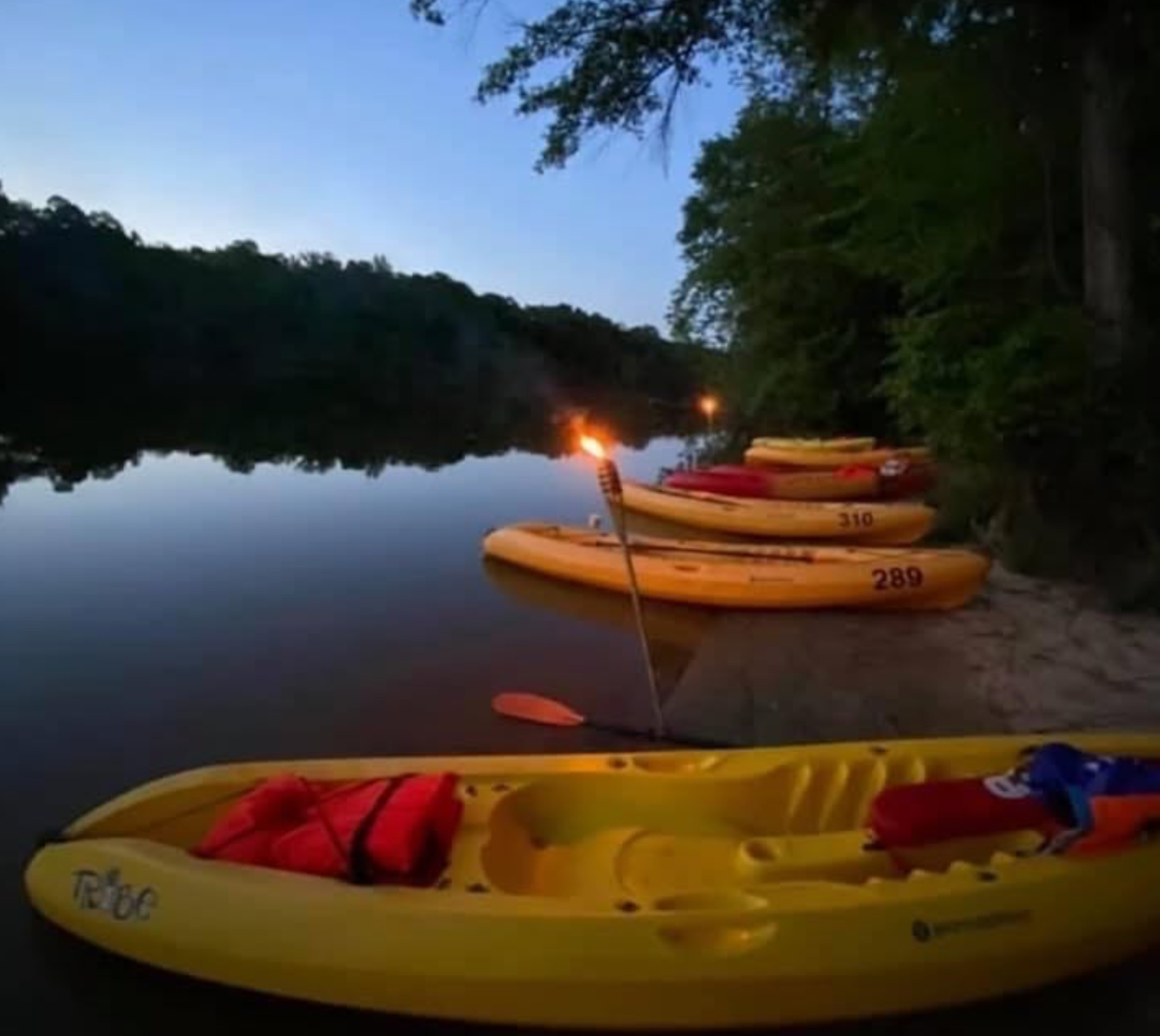 Yellow kayaks on a riverbank at dusk, lit torch, trees in background, calm water.