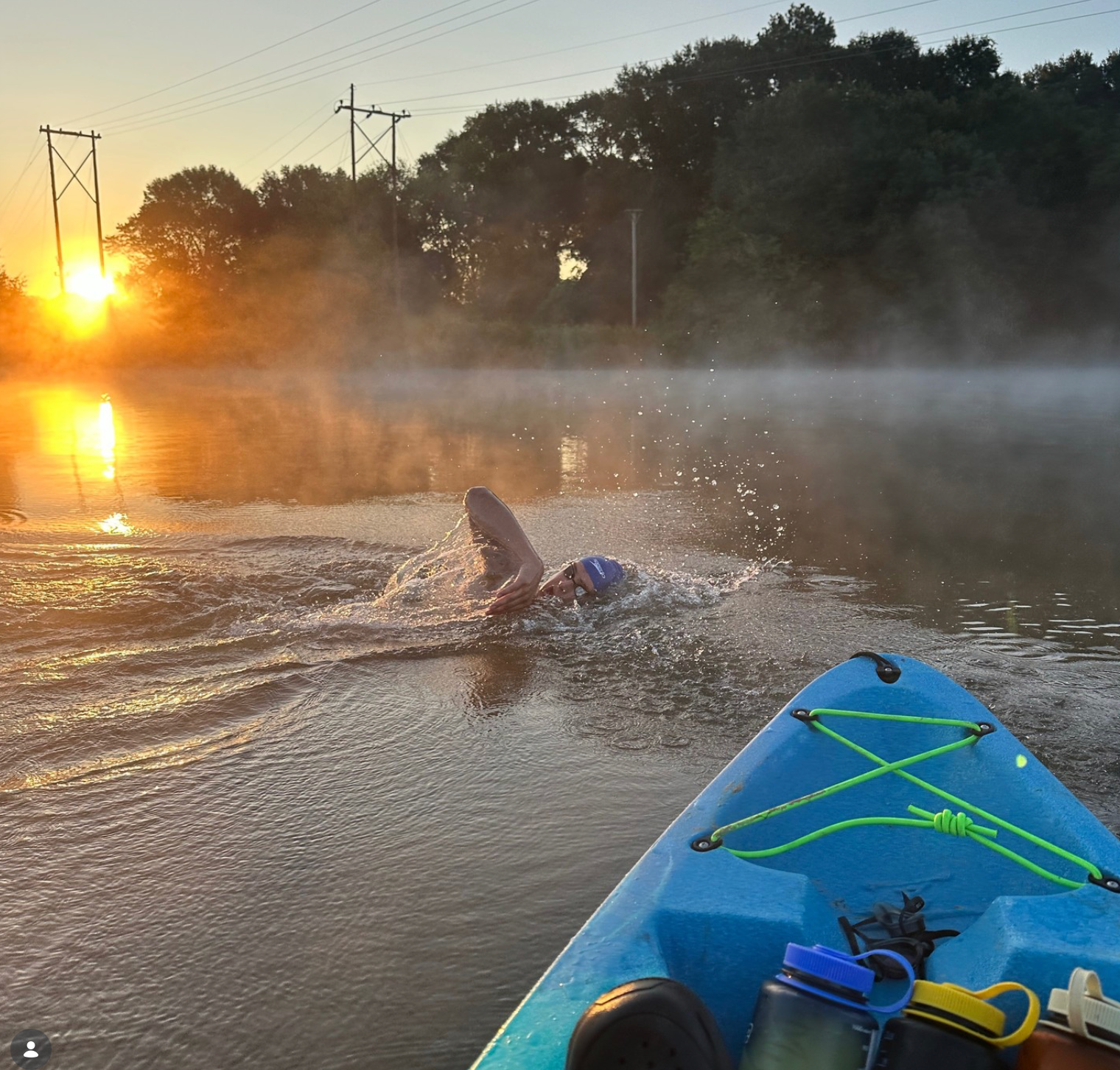 Swimmer in lake, kayak in foreground, sun rising, mist on water.