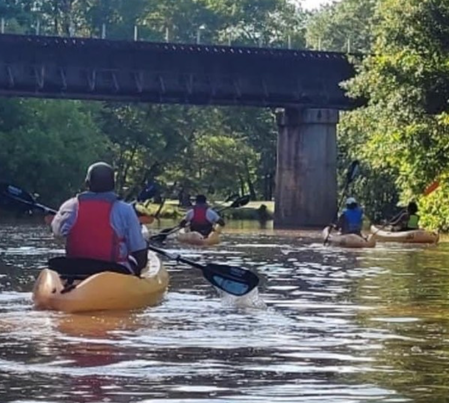 Kayakers paddling on a brown river under a bridge, wearing life vests.