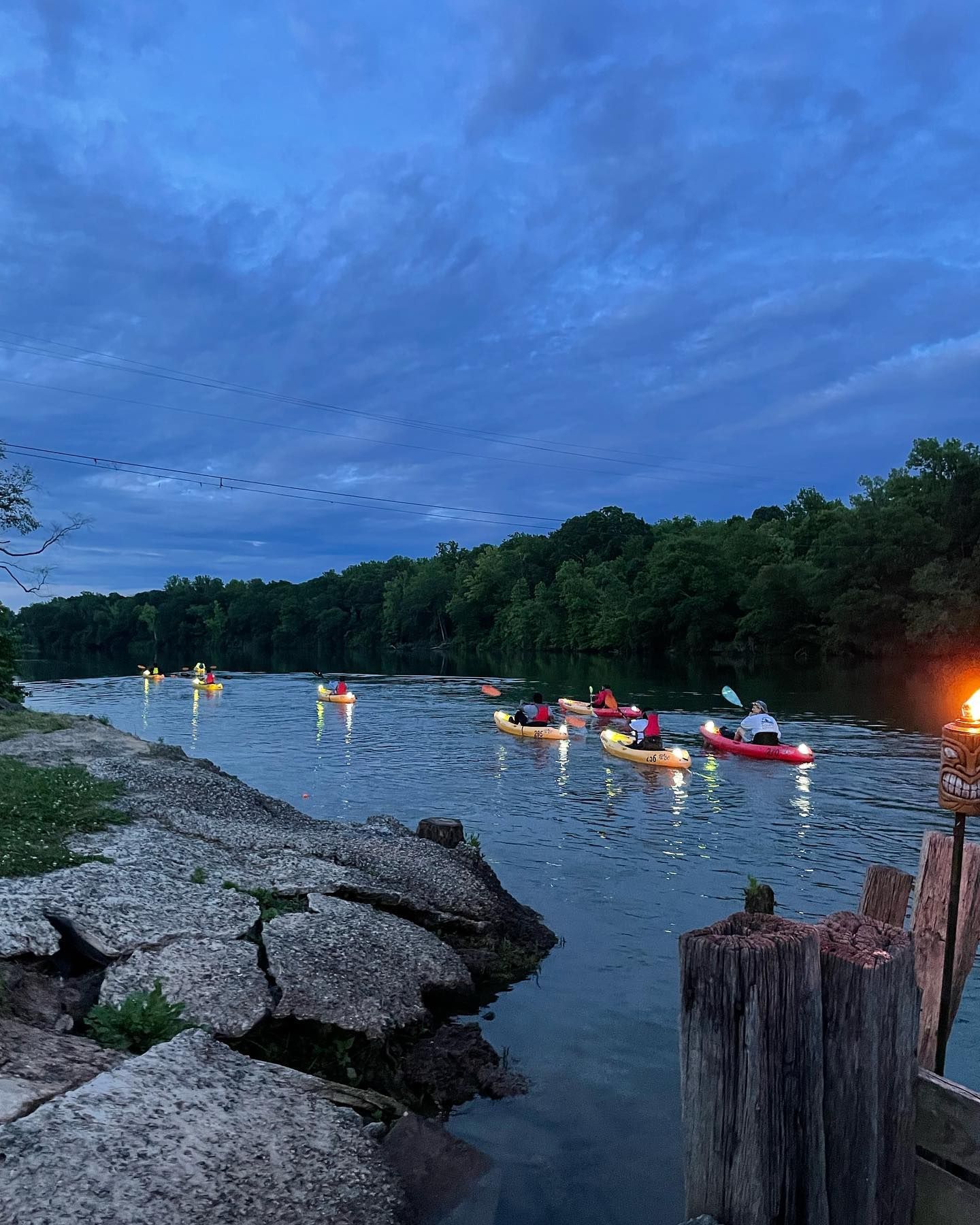 Kayakers with lights on a river at dusk, with trees along the banks.