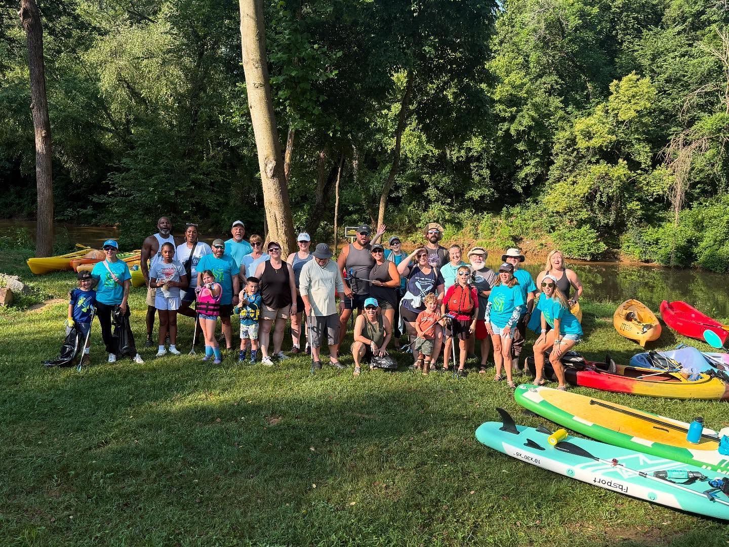 Group of people standing on grassy riverbank next to kayaks and paddleboards.