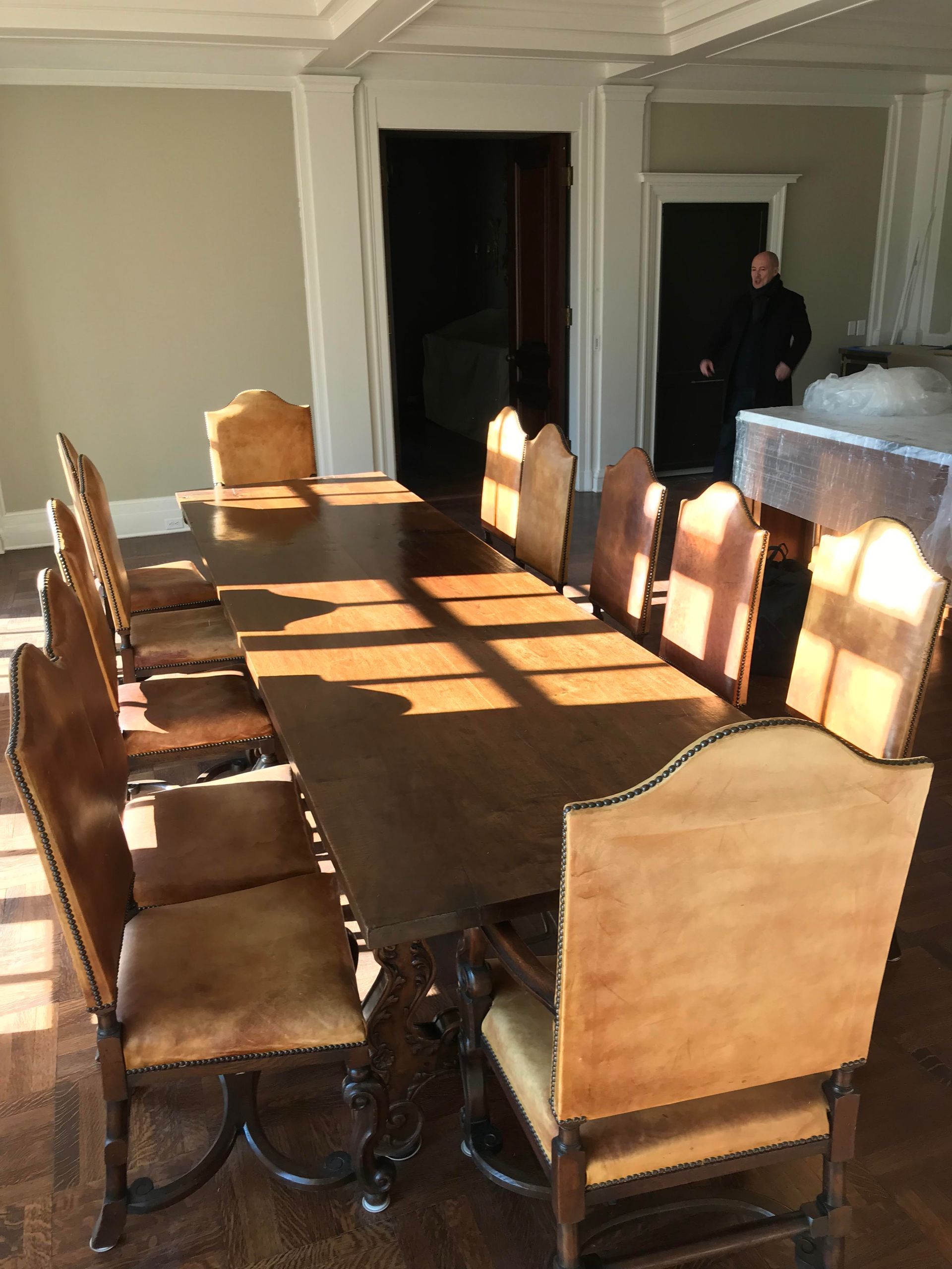 A long wooden dining table with 10 leather-backed chairs in a sunlit room. A person stands in a doorway in the background.