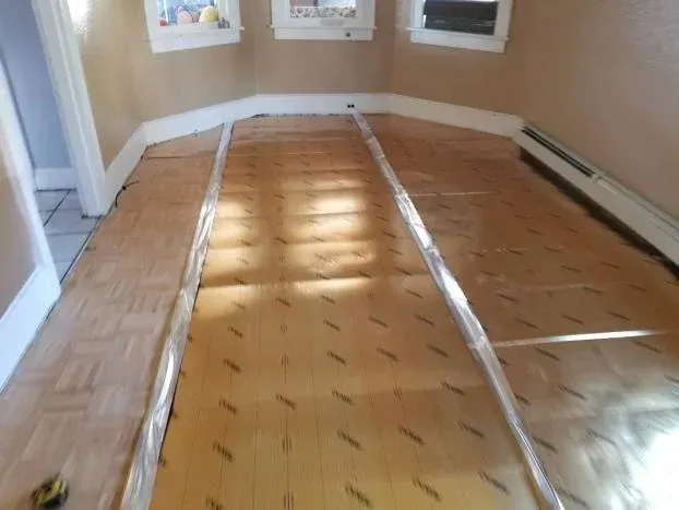 Room with parquet floor covered in paper. Two clear plastic tubes run lengthwise. Beige walls and trim.