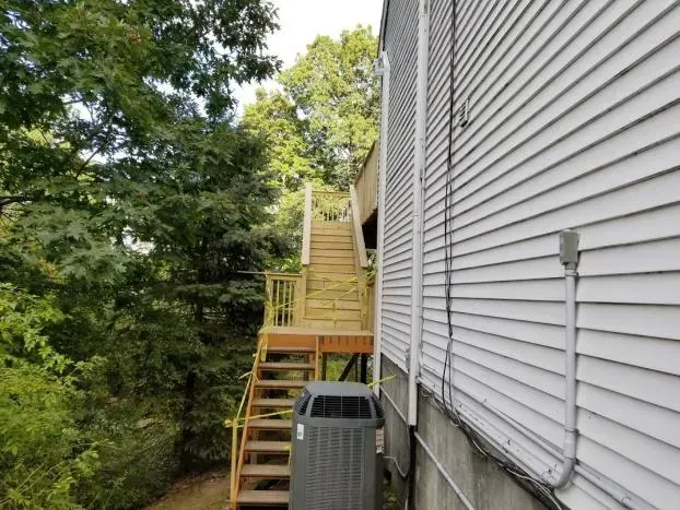Wooden staircase leading up from a deck, next to a white-sided building with an AC unit below.
