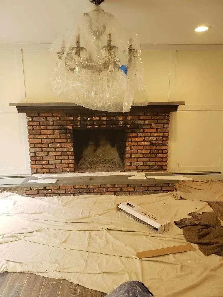 Brick fireplace covered in soot, with plastic-wrapped chandelier. Construction scene with drop cloths and tools.