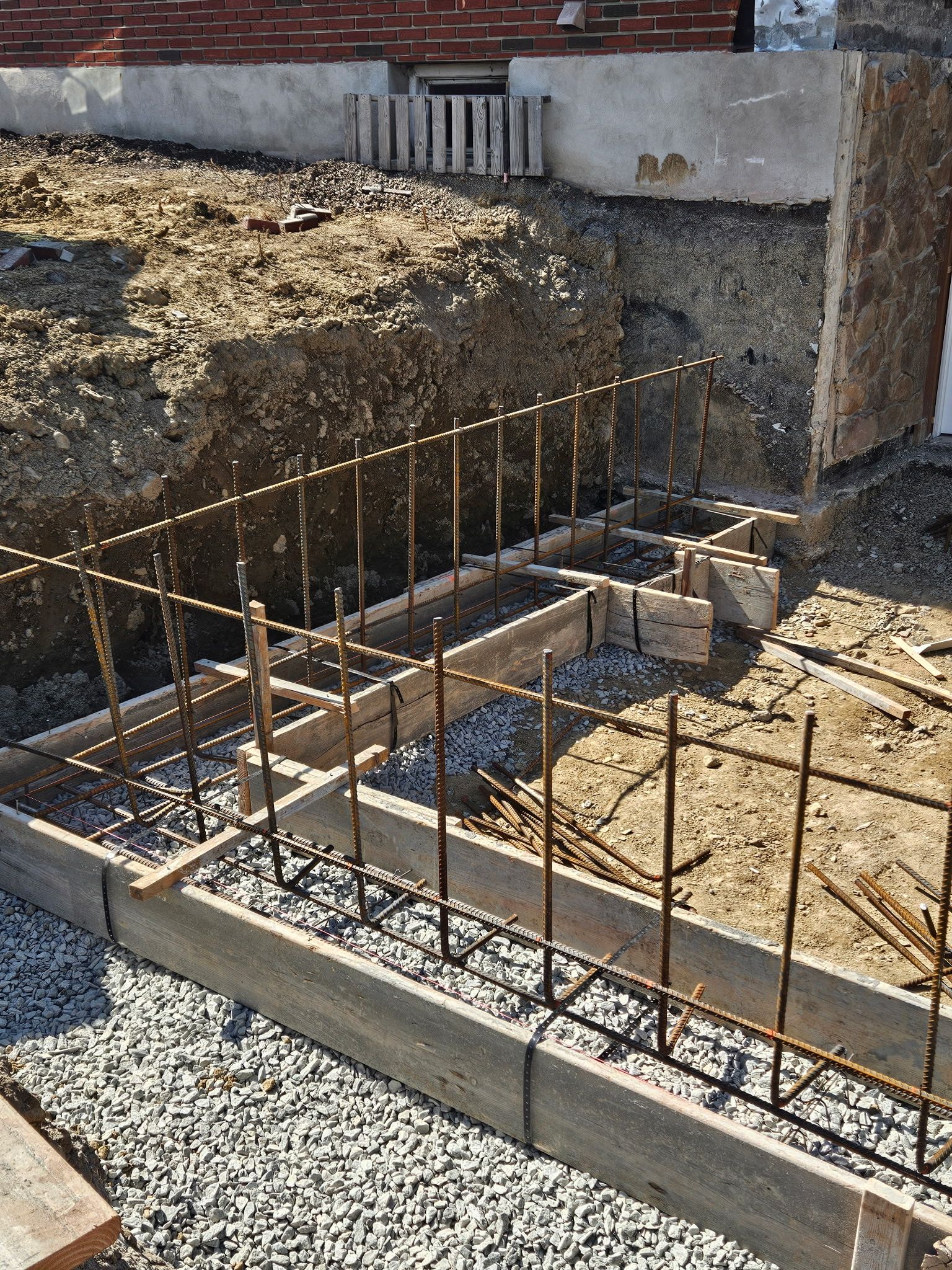 Foundation construction site, wooden forms with rebar, gravel base, dirt wall, brick building.
