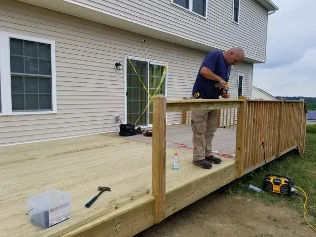 Man building a deck on a house. He's using a drill, with tools and supplies around him, outside.