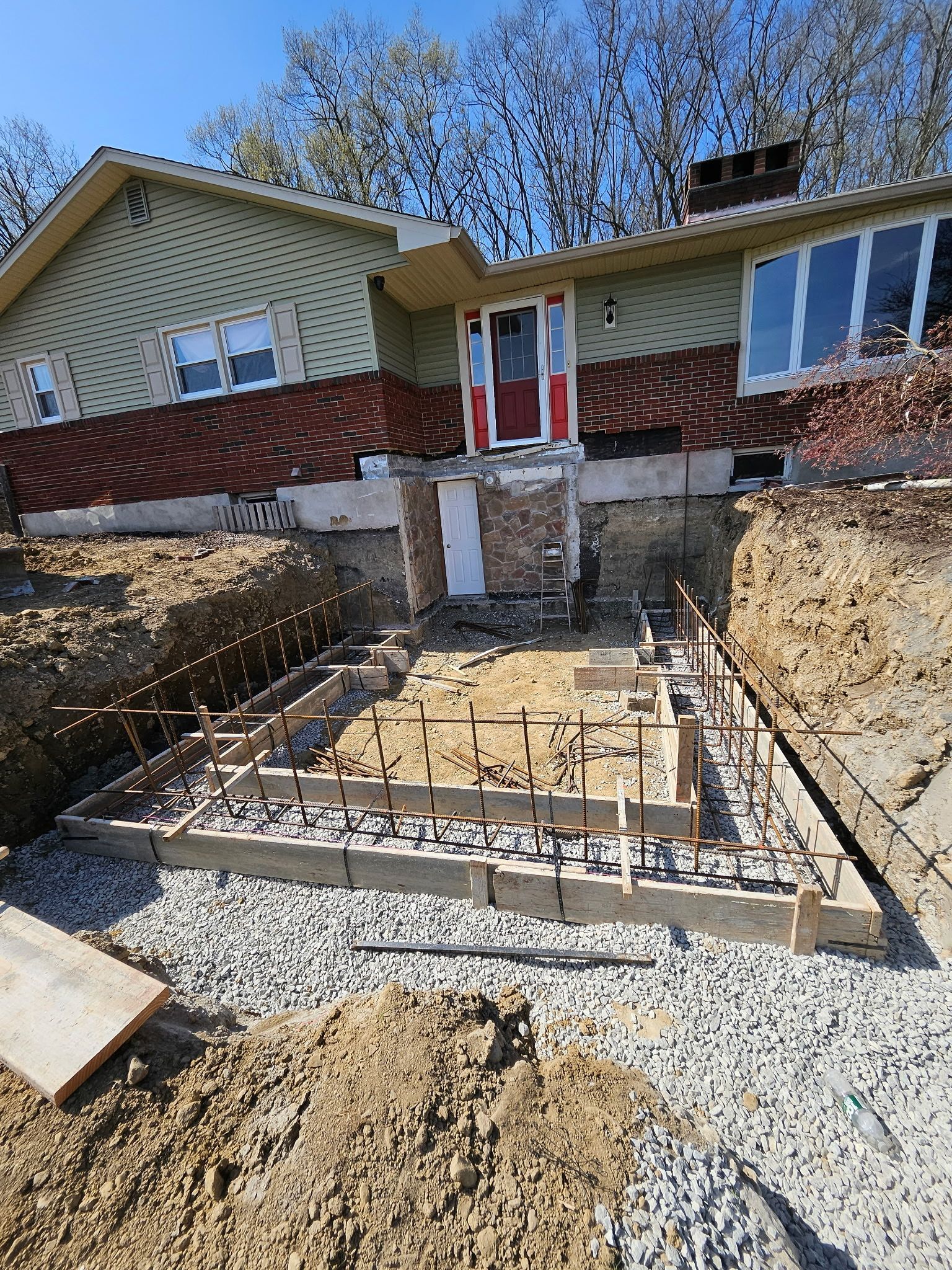Foundation construction in progress at a house. Wooden forms, rebar, and gravel visible in excavated area.