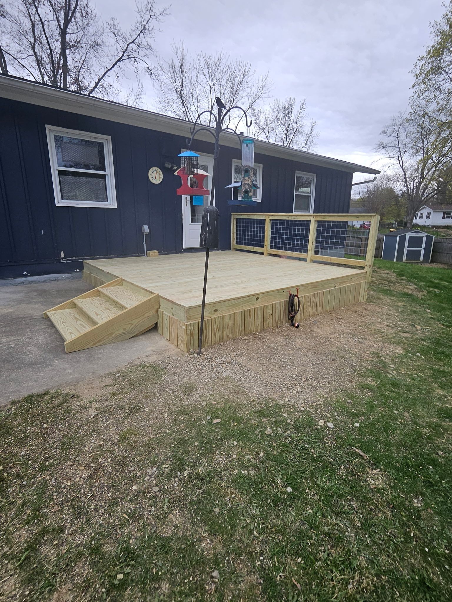 Newly constructed wooden deck outside a dark blue house with steps leading up to it; bird feeder in foreground.