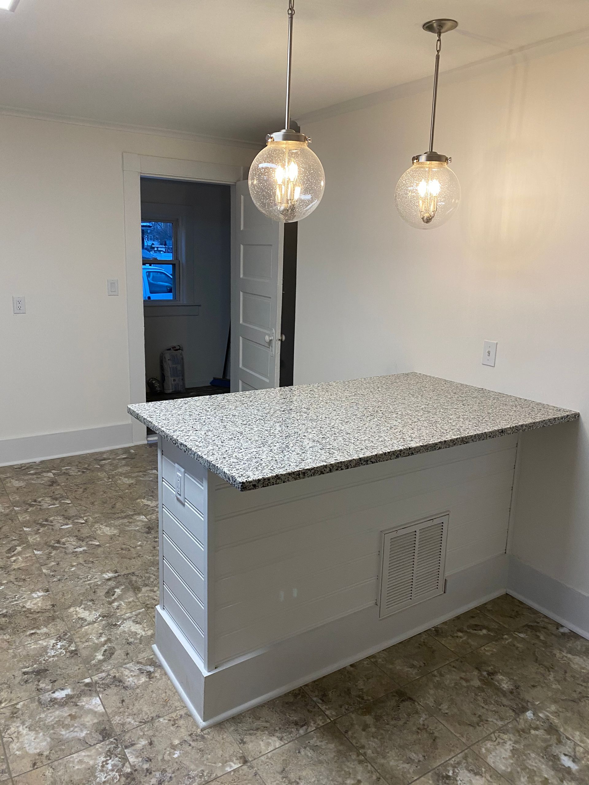 A white kitchen island with granite countertop, two pendant lights, and an open doorway in a room.