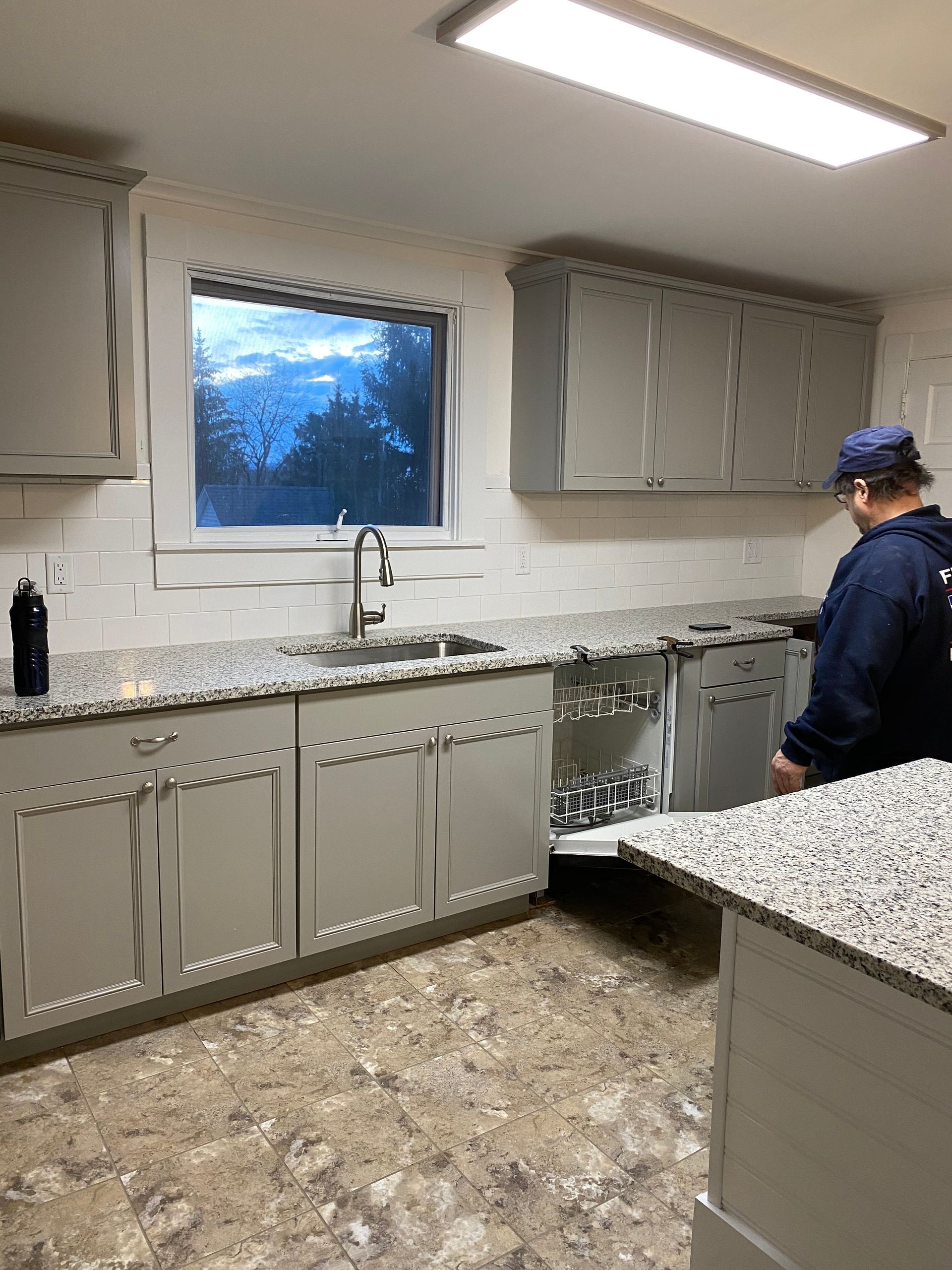 Kitchen with gray cabinets, granite countertops, a person, and a window with a cloudy sky.