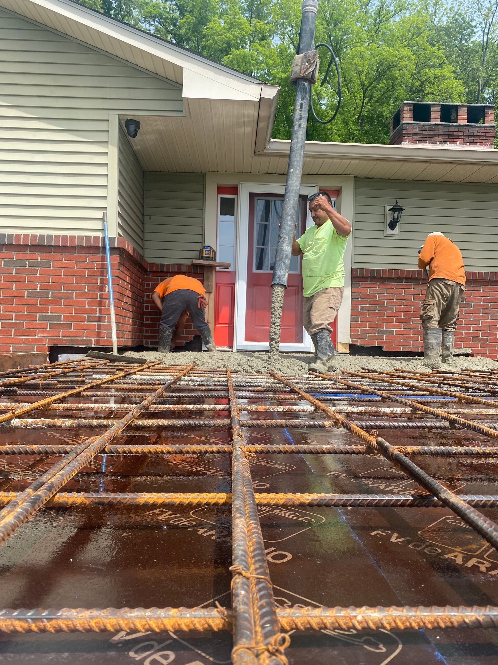 Construction workers pouring concrete onto rebar grid, next to a brick house, on a sunny day.