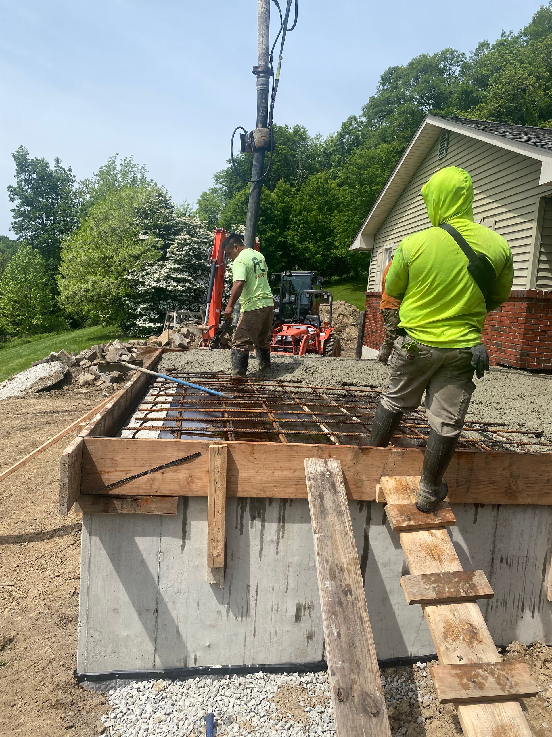 Construction workers pouring concrete for a foundation with a pump truck. Green safety vests, sunny day.
