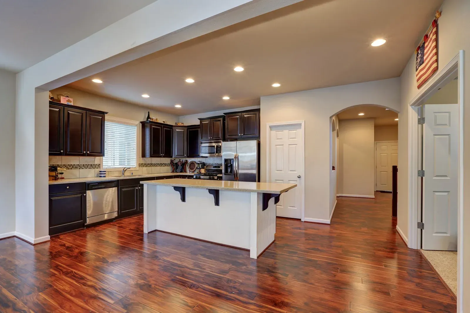 Open-concept kitchen with dark cabinets, white island, and hardwood floors; an archway leads to a hallway.