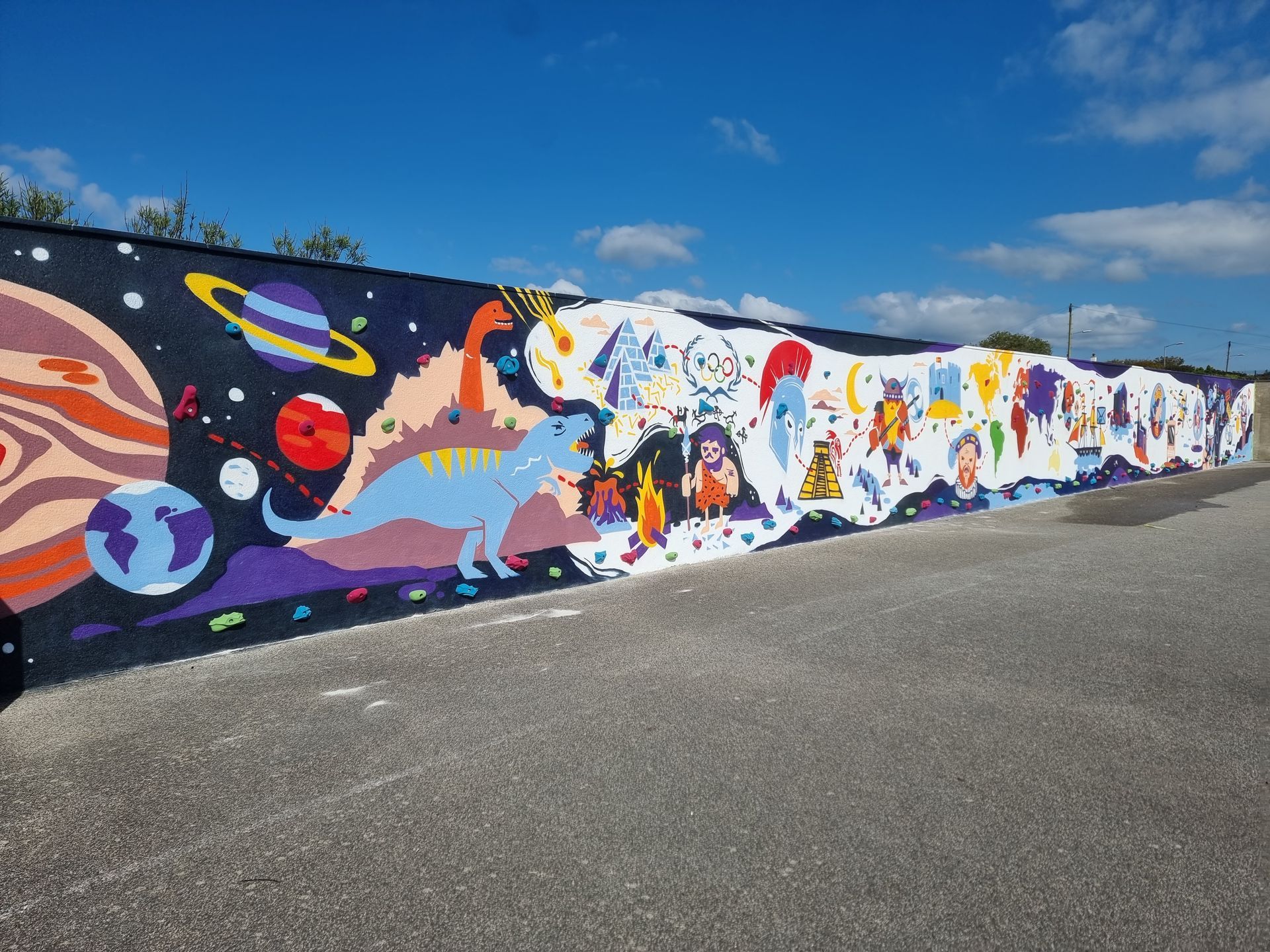 Mural: Planets, dinosaur, and colorful cartoon figures against a starry sky on a long wall under blue sky.