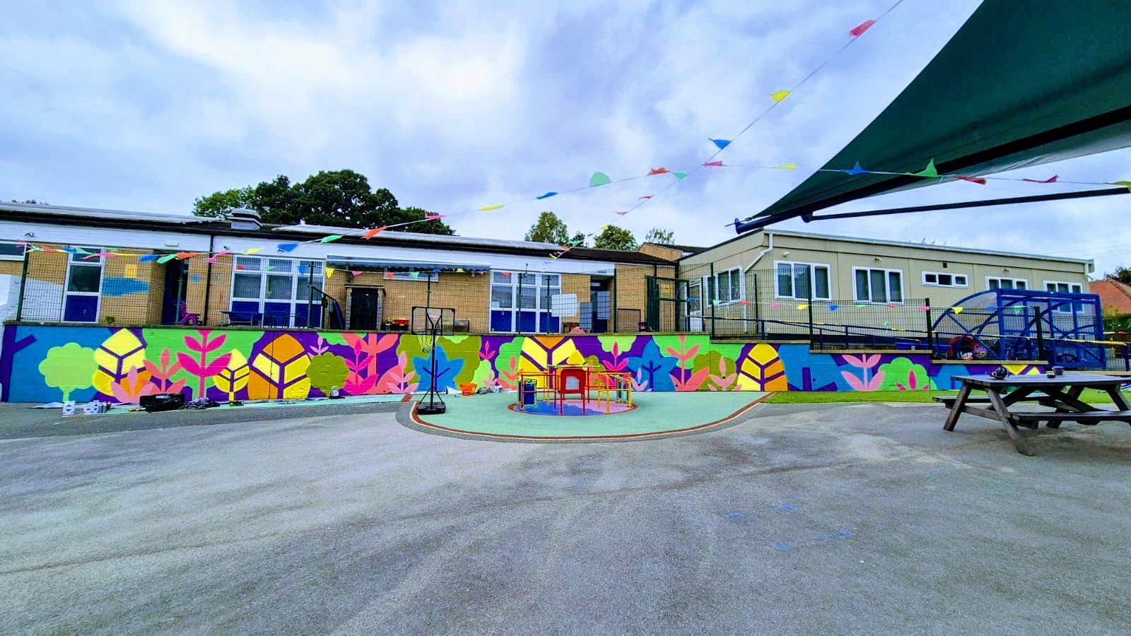 Colorful mural on a building's exterior wall with a play area in front; blue sky, festive flags, and a canopy.