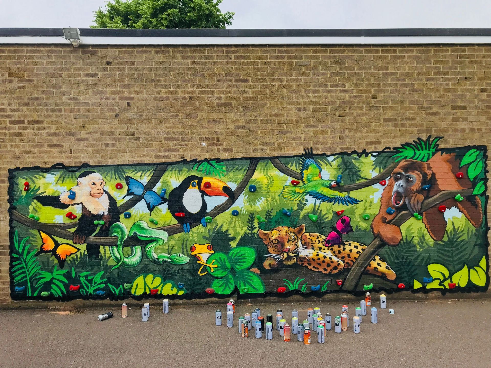 Mural of a jungle scene with monkeys, birds, and a leopard on a brick wall, surrounded by spray paint cans.