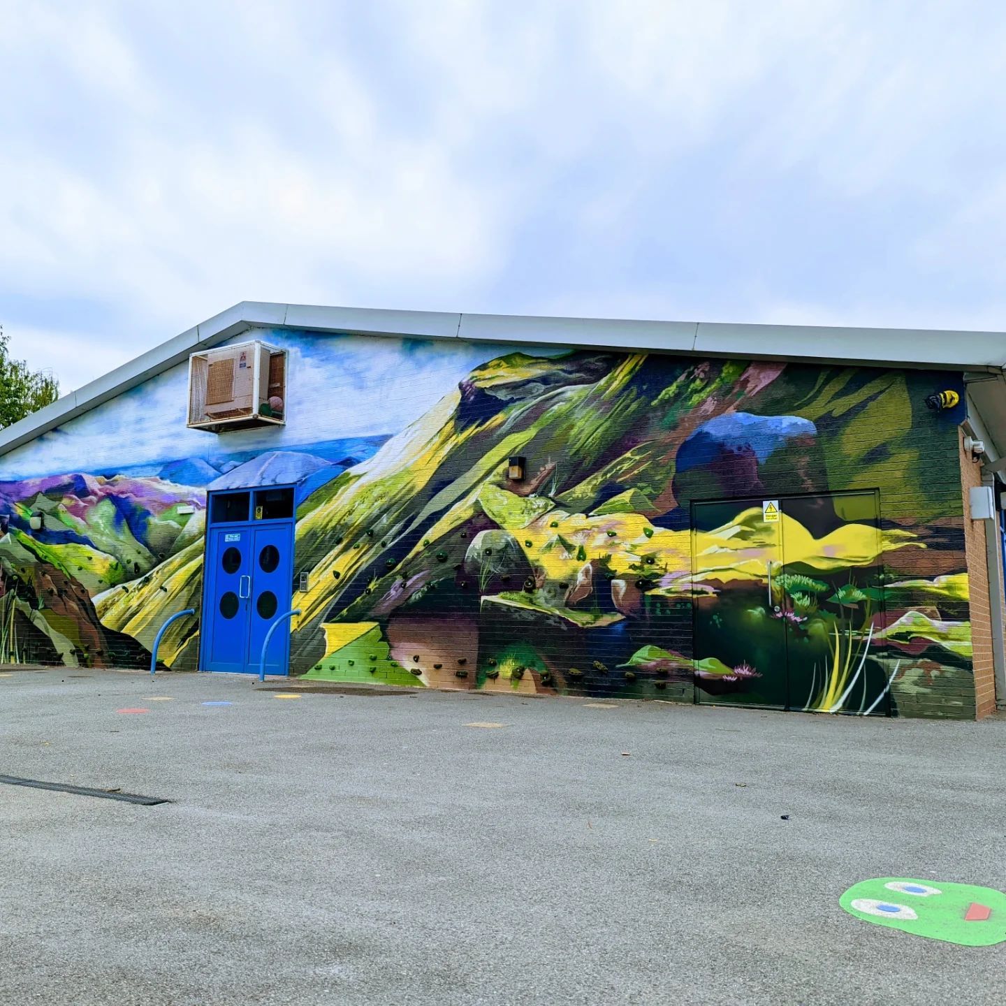 Mural of a mountain landscape on a blue-doored building with an AC unit; gray pavement in the foreground.