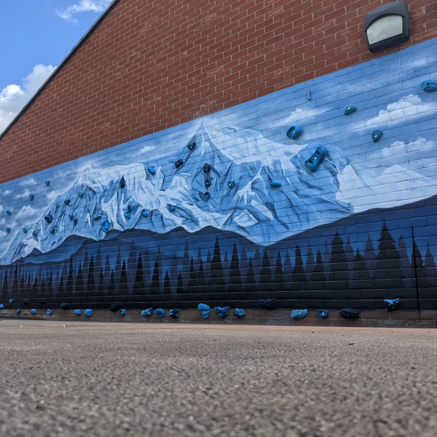 Mural on brick wall depicts a blue and white mountain range above a dark forest; climbing holds are affixed.