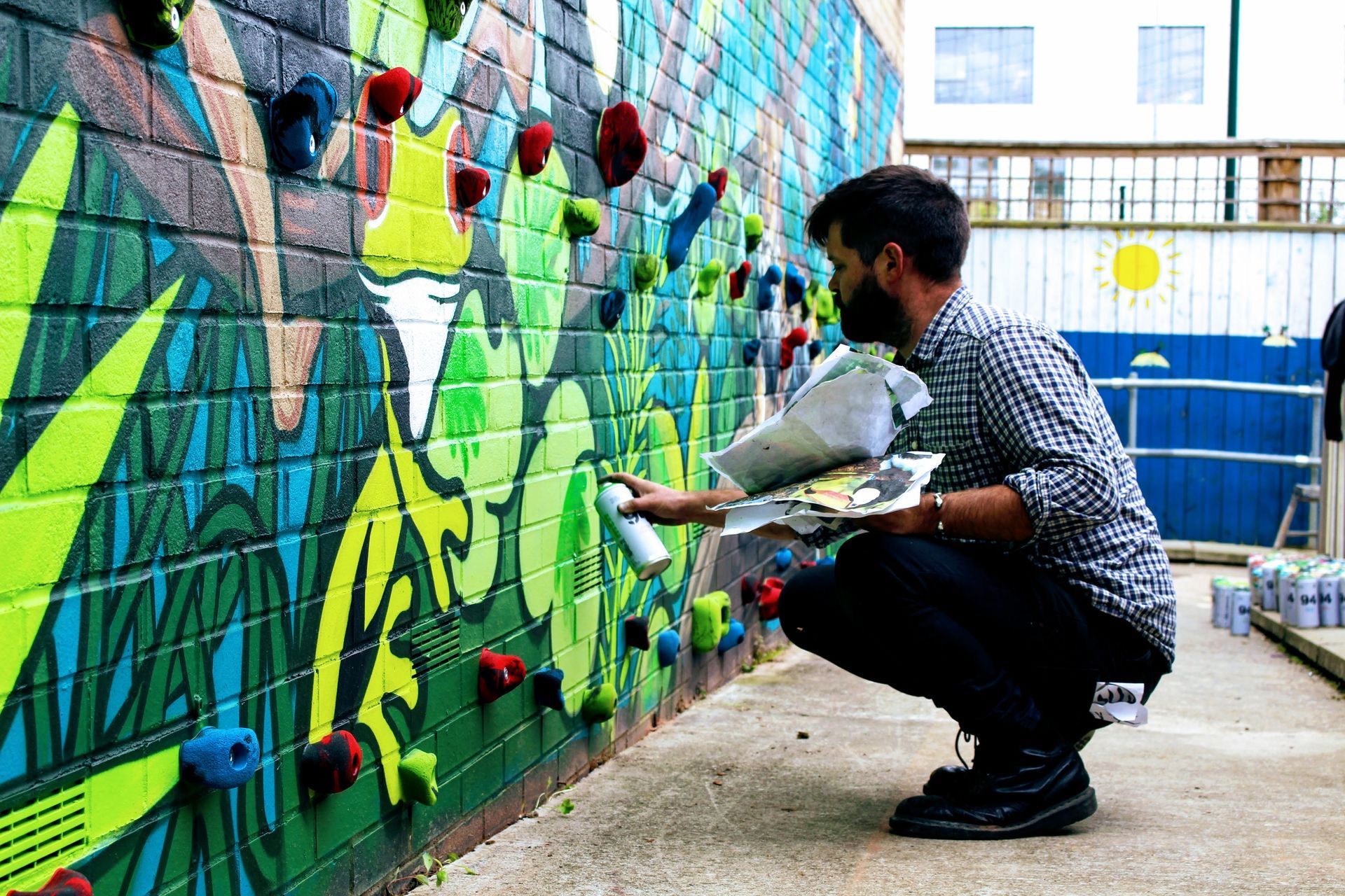 Man spray paints a colorful mural on a brick wall with climbing holds.
