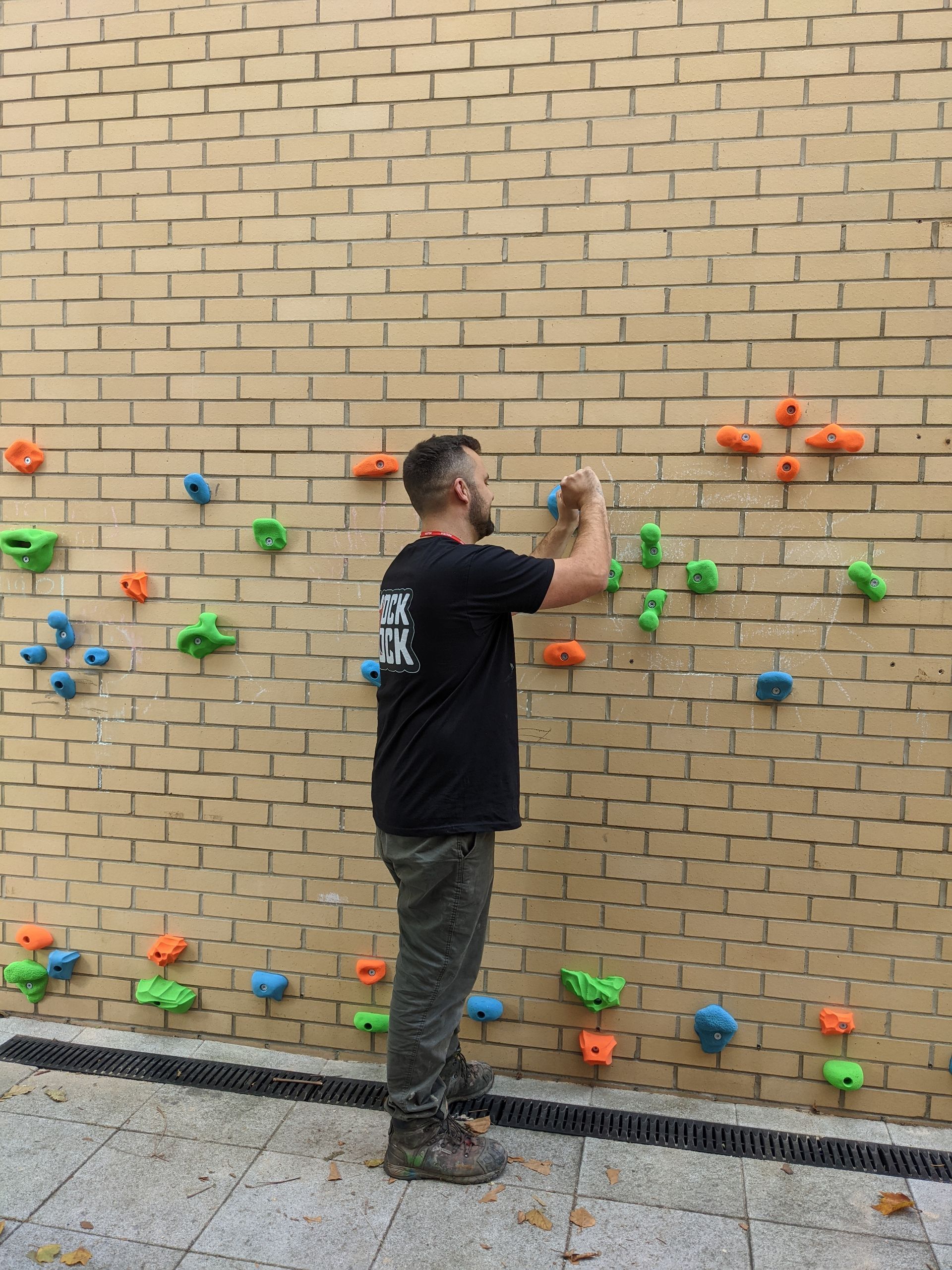 Man on a climbing wall, reaching for a hold. Brick wall with colorful handholds. Outdoors.