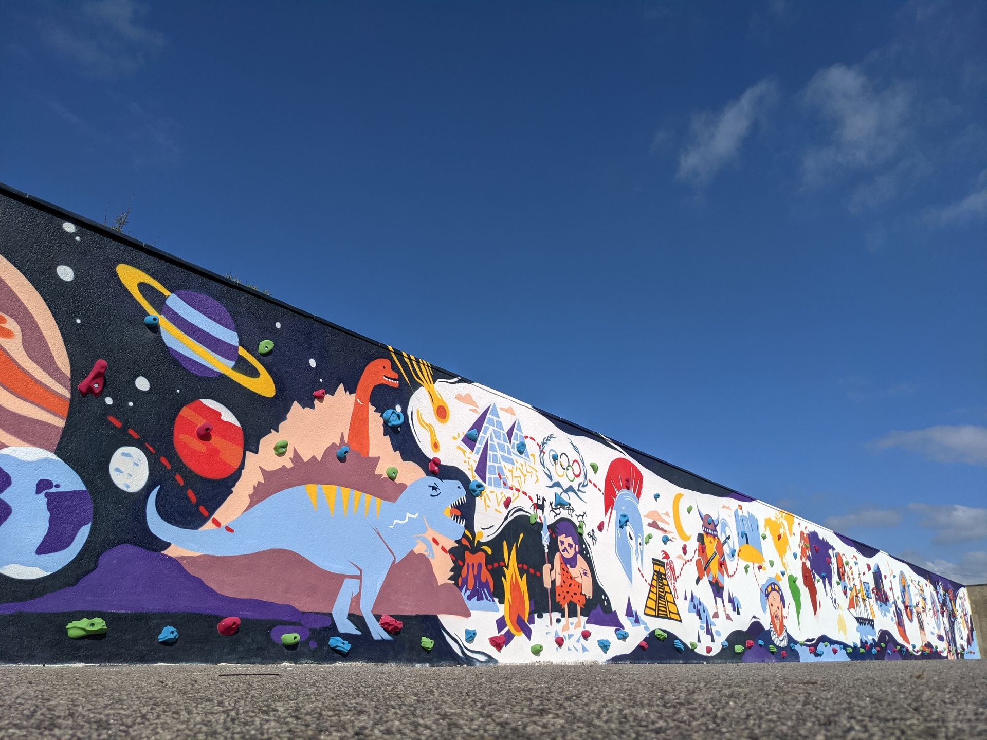 Mural of planets, a dinosaur, and colorful creatures against a blue sky, painted on a long wall.