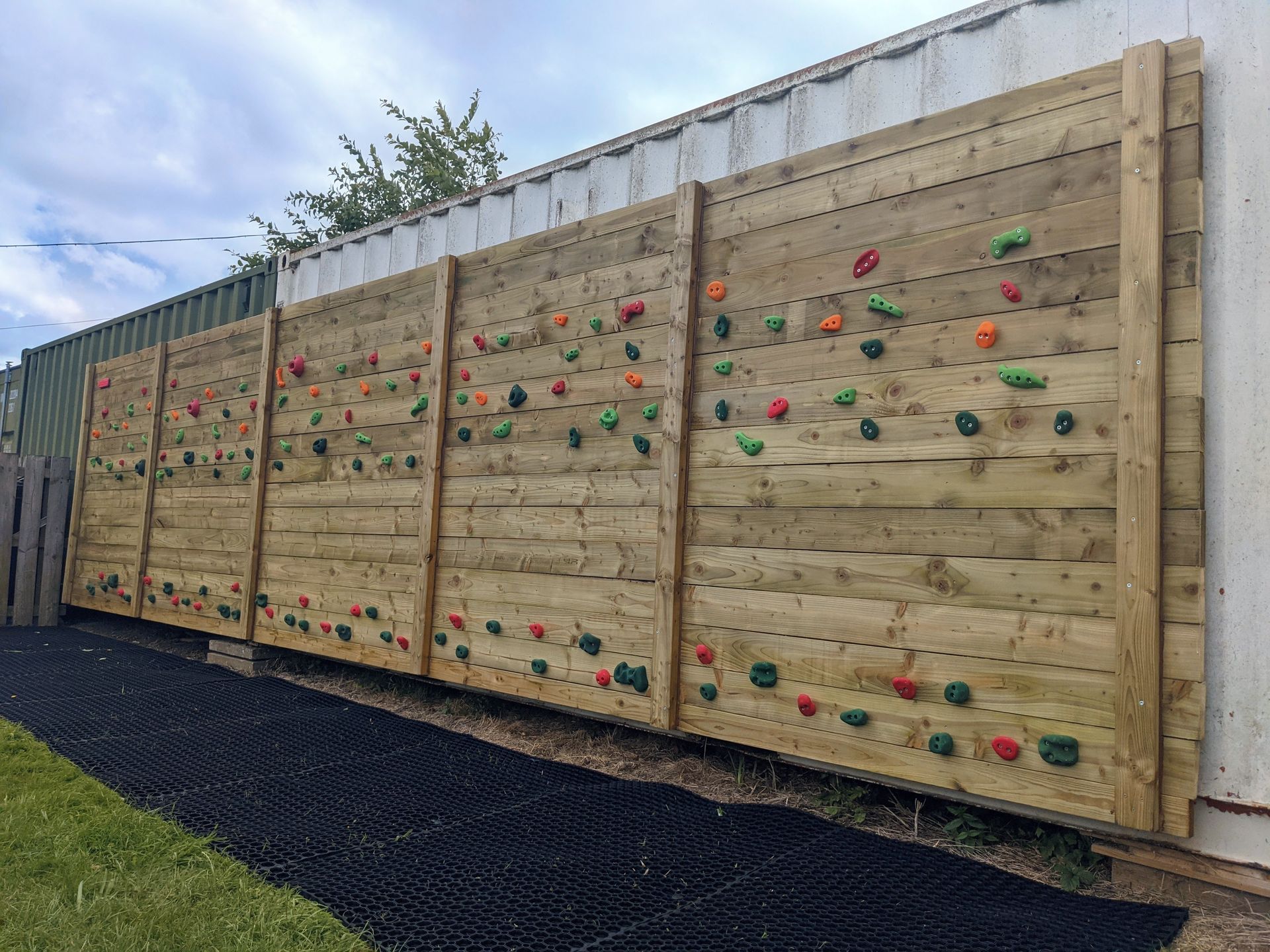 Wooden outdoor climbing wall with colored holds against a white building and blue sky.