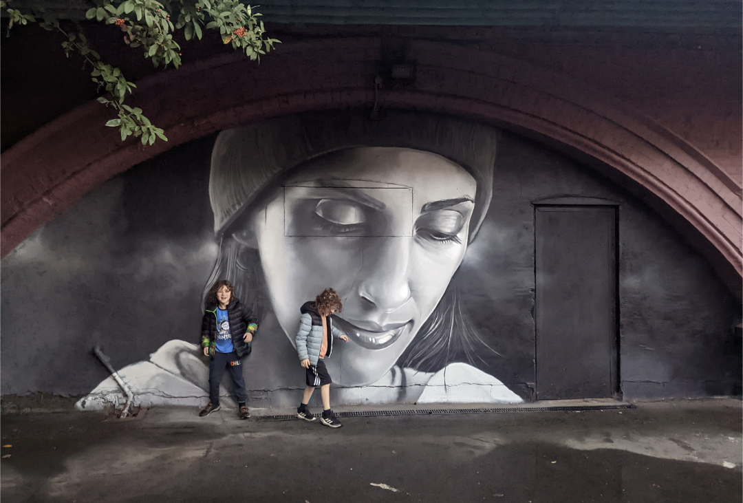 Two children stand before a large black-and-white mural of a woman's face under an archway.