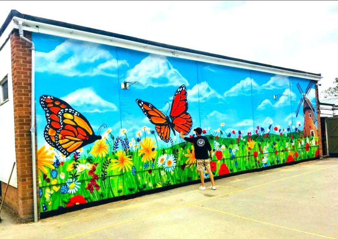 Colorful mural of butterflies, flowers, windmill, and blue sky painted on a building exterior. Person standing next to the mural.