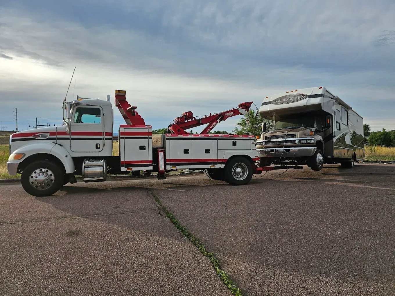 Tow truck towing a damaged RV on an asphalt road under a cloudy sky. The truck is white and red.