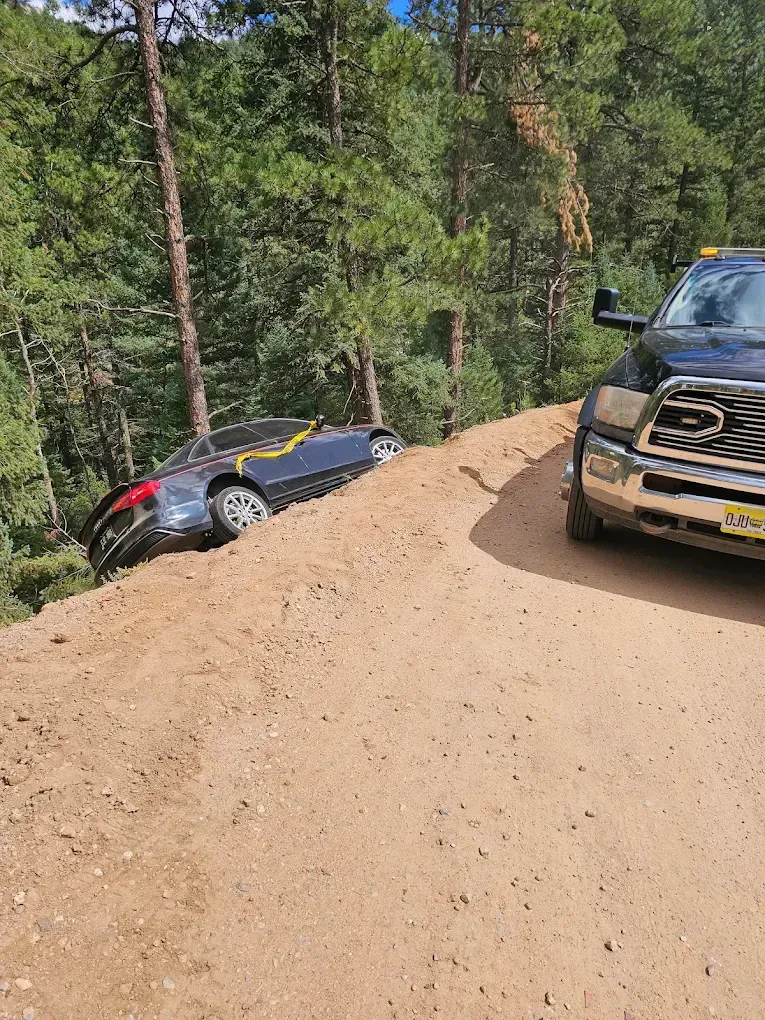 Dark car on dirt hillside next to tow truck on a dirt road in a wooded area.
