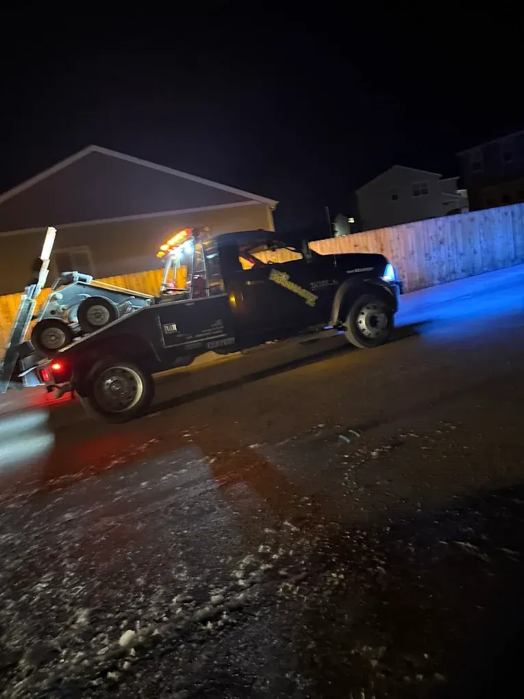 Black tow truck towing a car at night. Bright lights illuminate the vehicles and snow-covered road.
