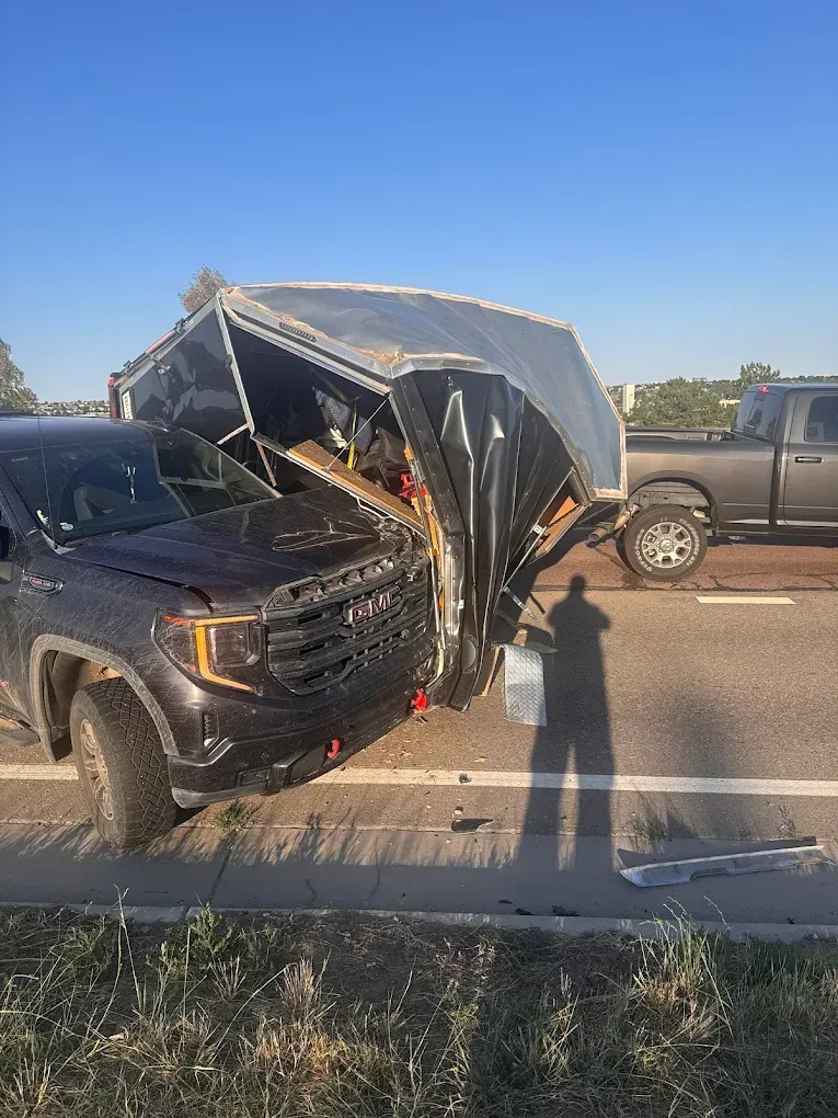 Black truck with damaged front collided with a cargo trailer. Another truck is parked behind them.