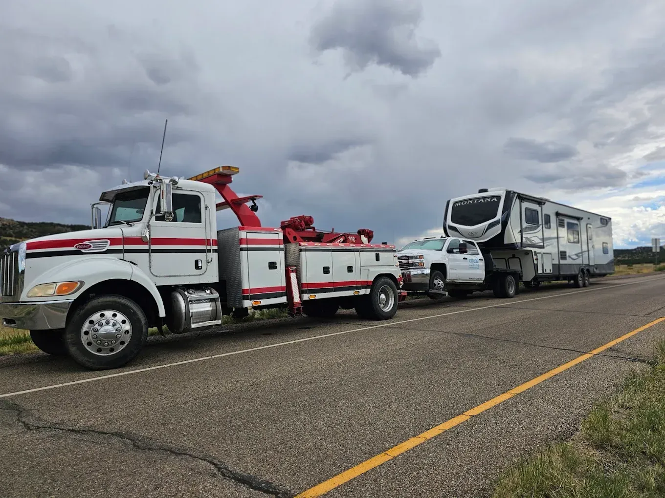 Tow truck towing a pickup truck hauling a fifth-wheel RV on a road under a cloudy sky.