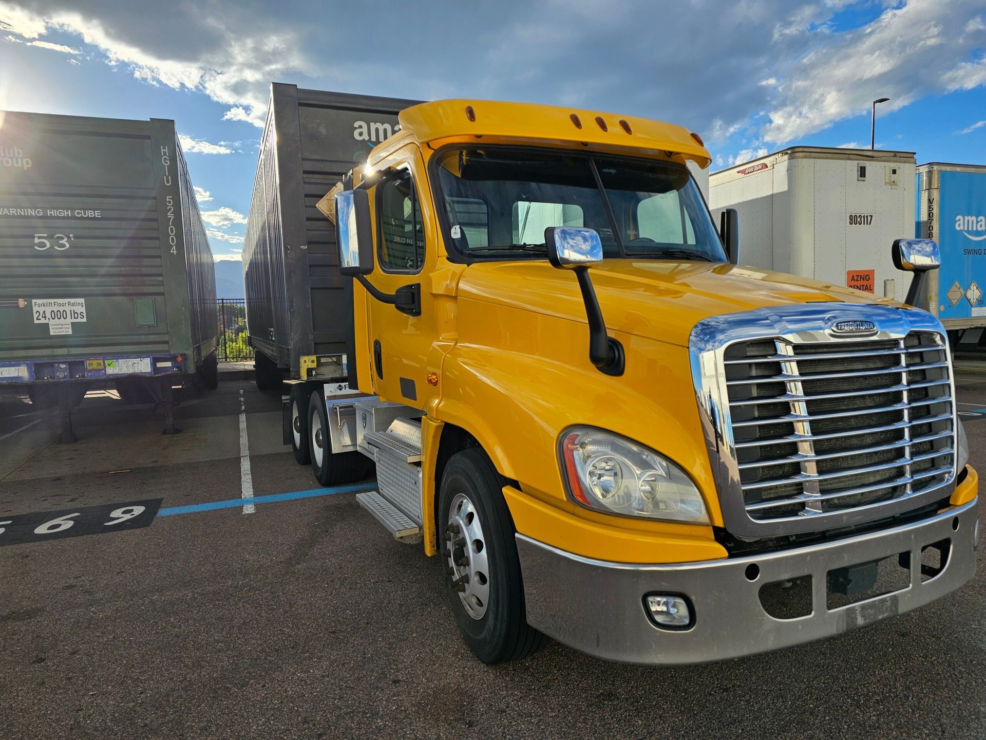 Yellow semi-truck with Amazon trailer parked in a lot, cloudy sky in the background.