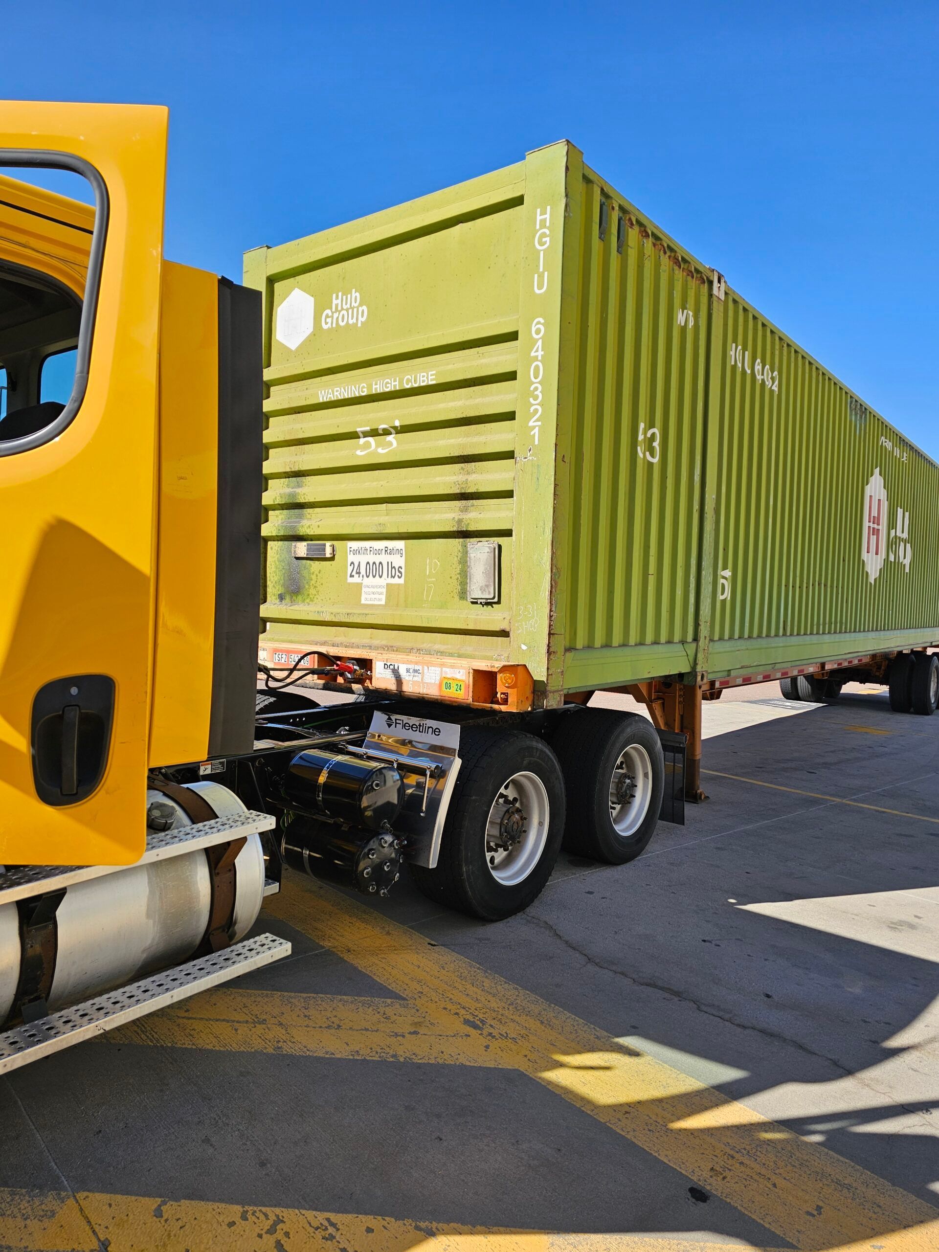 Yellow semi-truck with green shipping container on a sunny day.