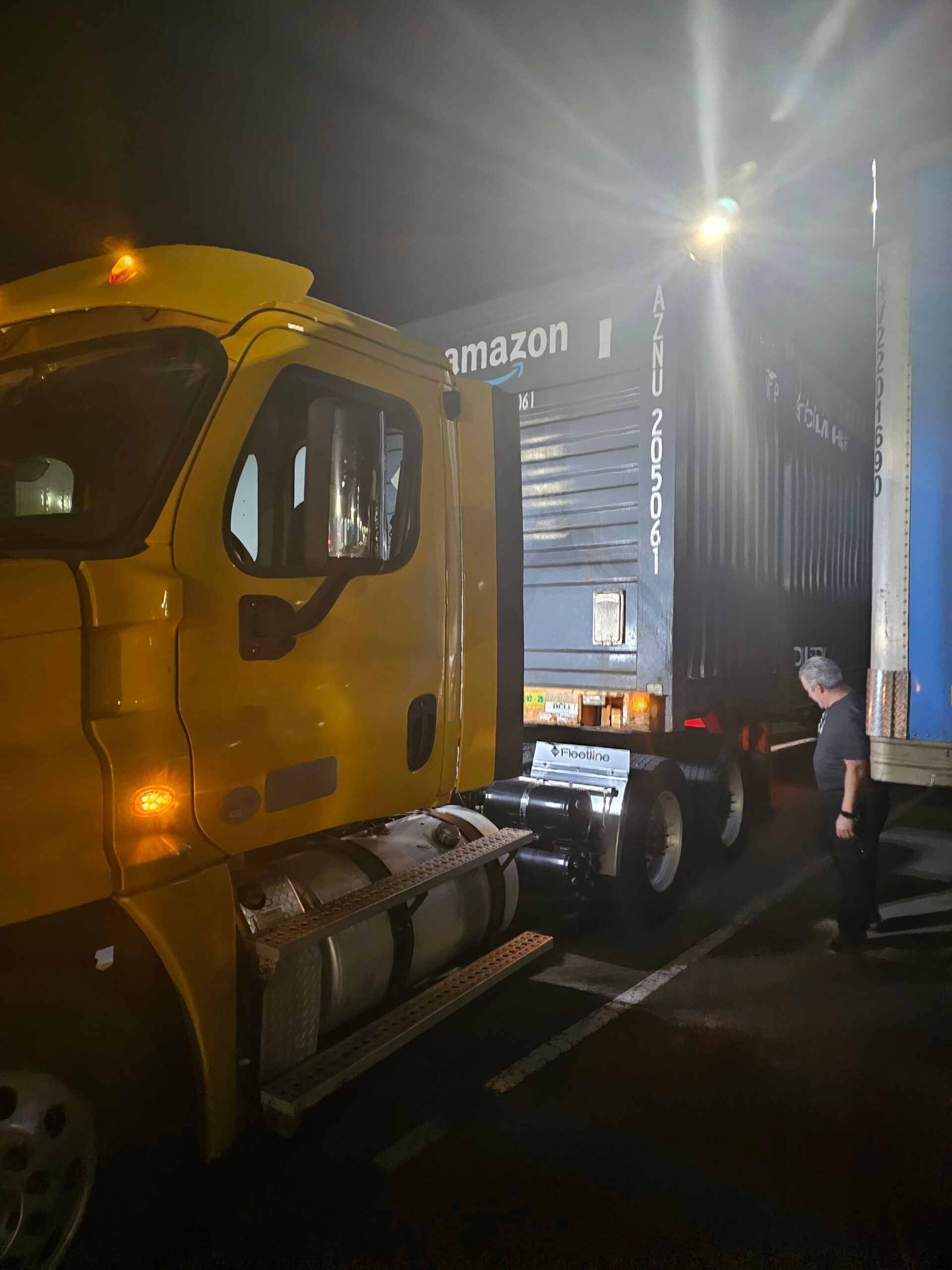 Yellow Amazon truck next to a shipping container; a person looks at the container's interior. Dark setting.