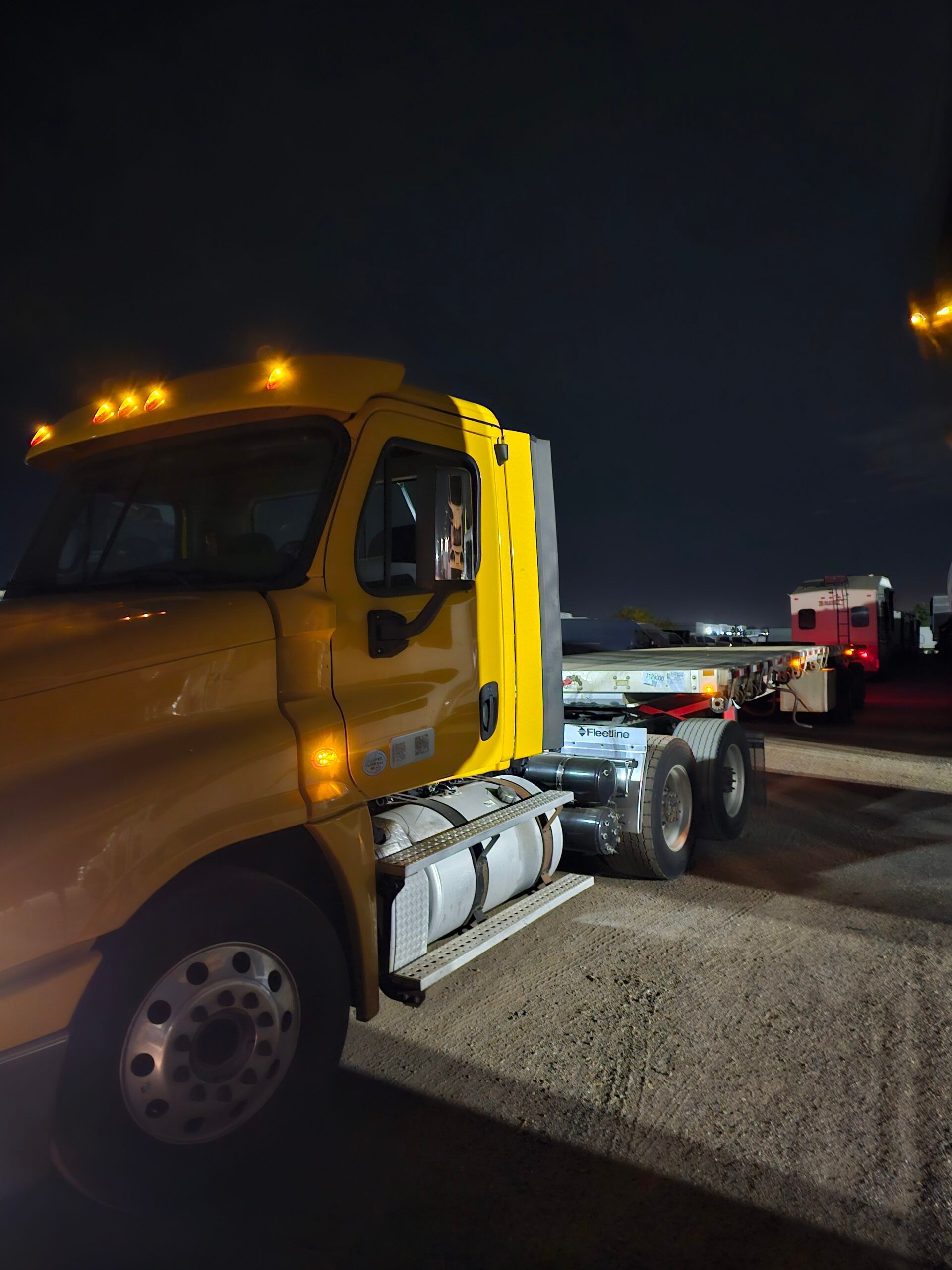Yellow semi-truck with flatbed trailer at night, headlights on.