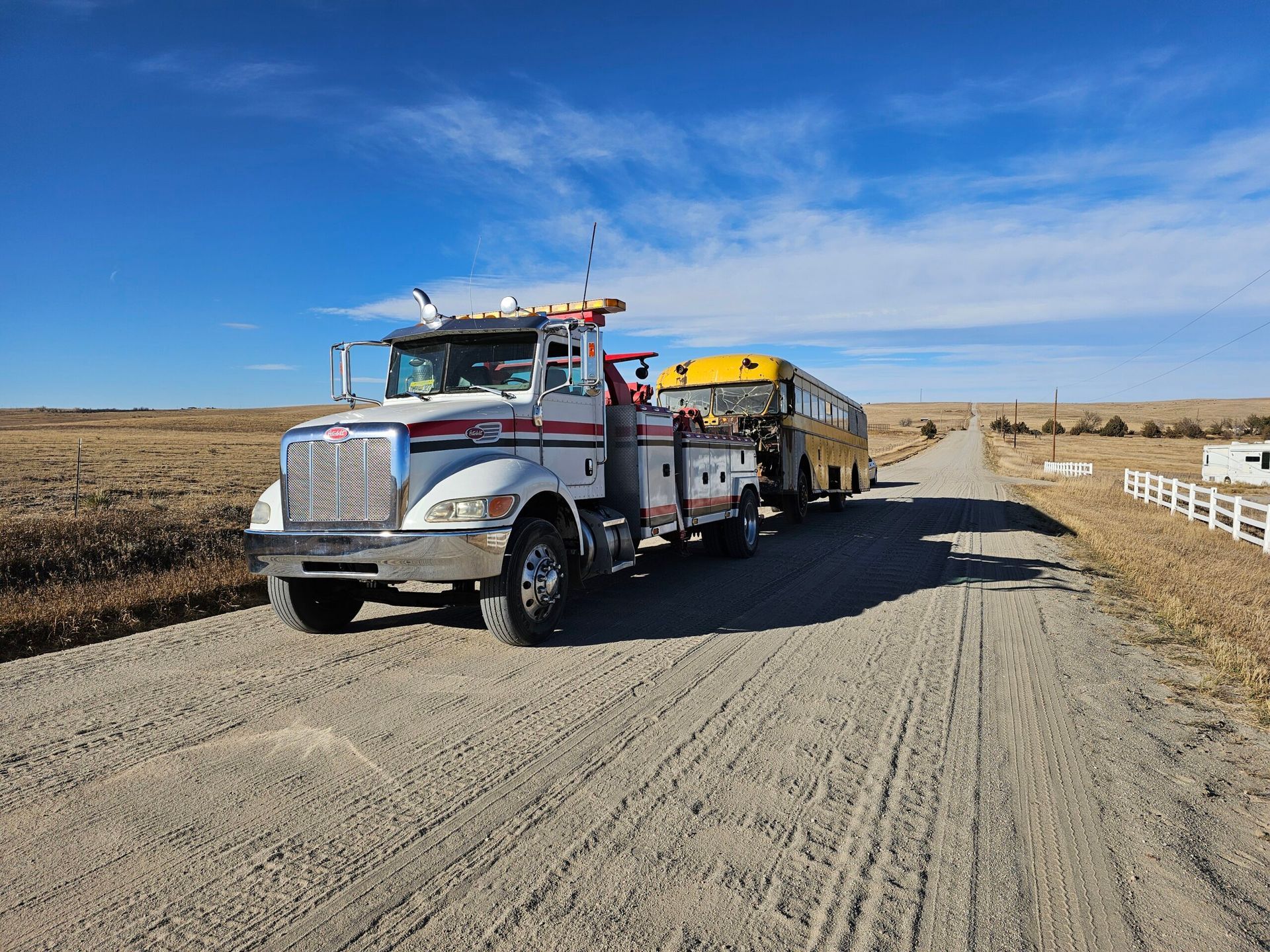 Tow truck hauling a yellow school bus on a dirt road under a clear blue sky.