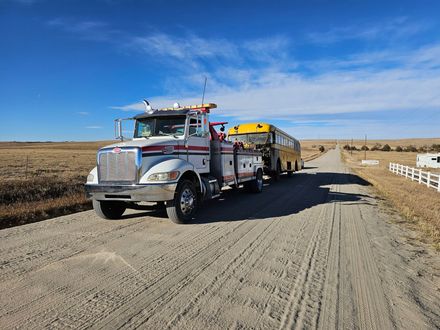 Tow truck towing a yellow school bus on a dirt road under a blue sky in a rural setting.