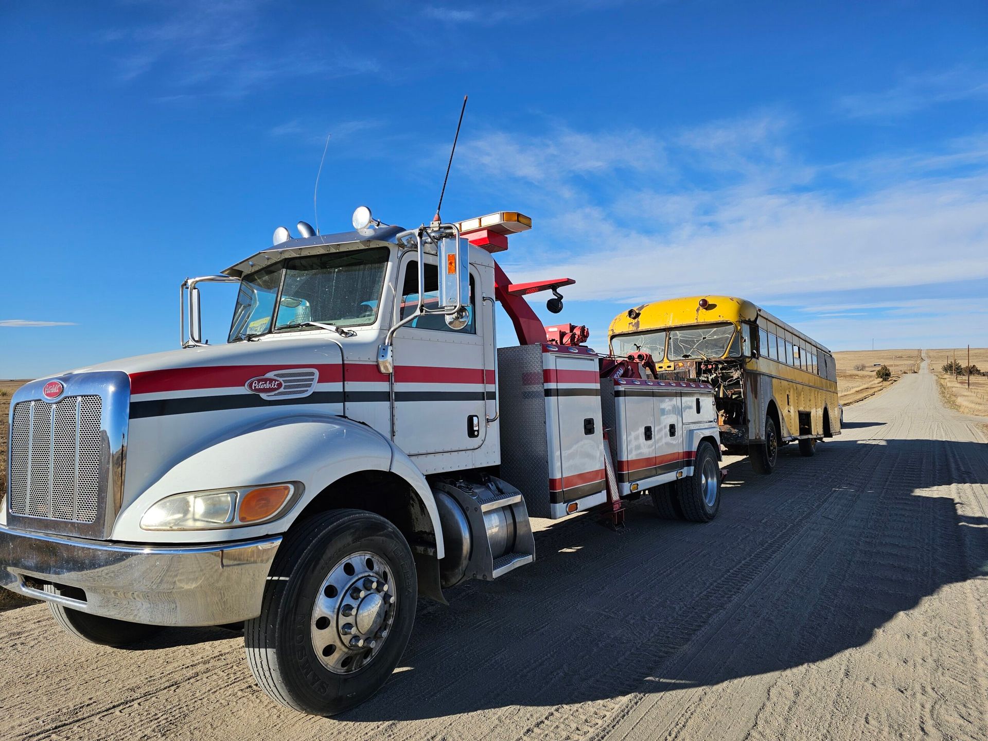 Tow truck towing a yellow school bus on a dirt road under a blue sky.
