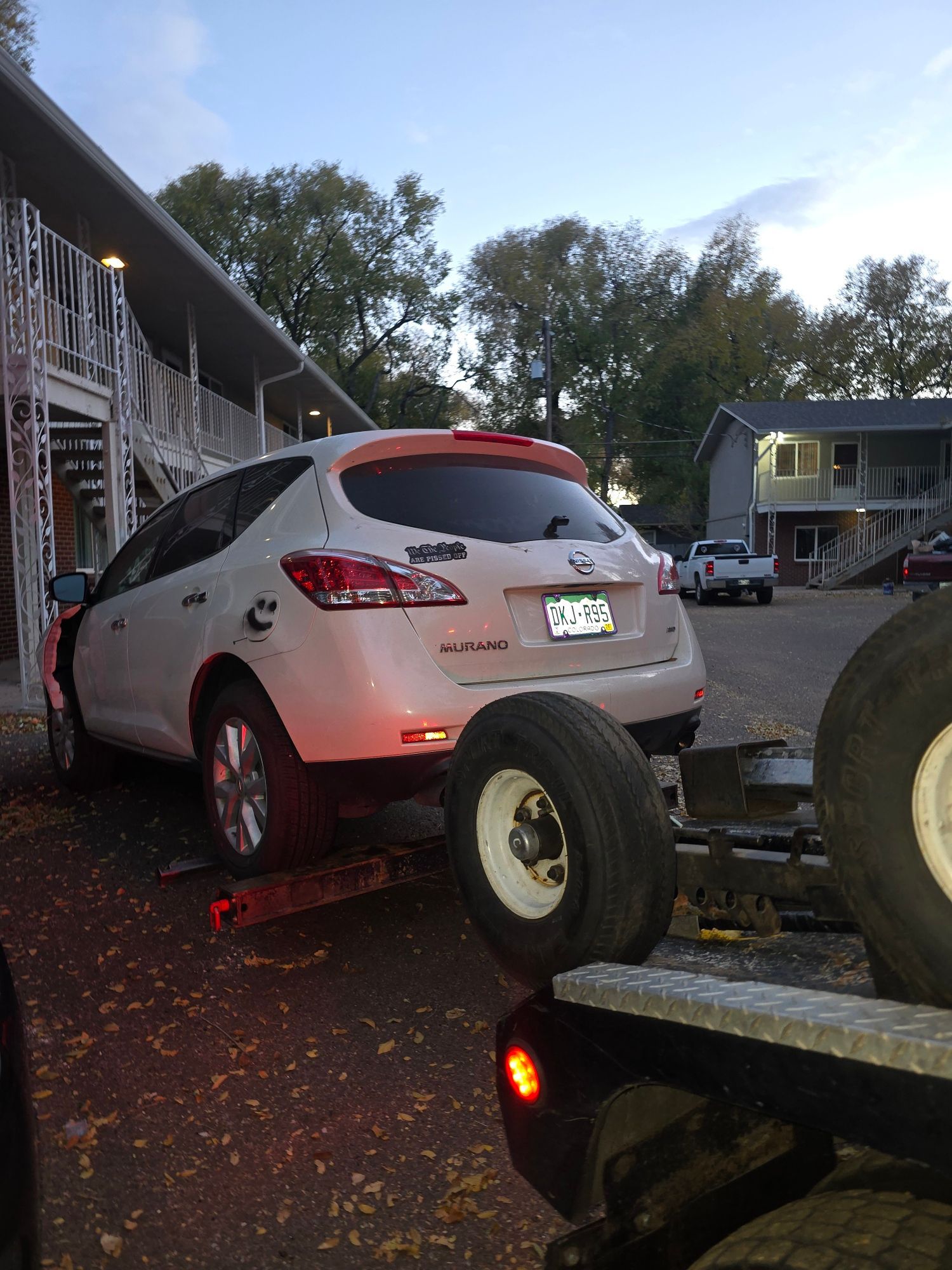 White SUV being towed onto a flatbed trailer, parked near a building with stairs, at dusk.