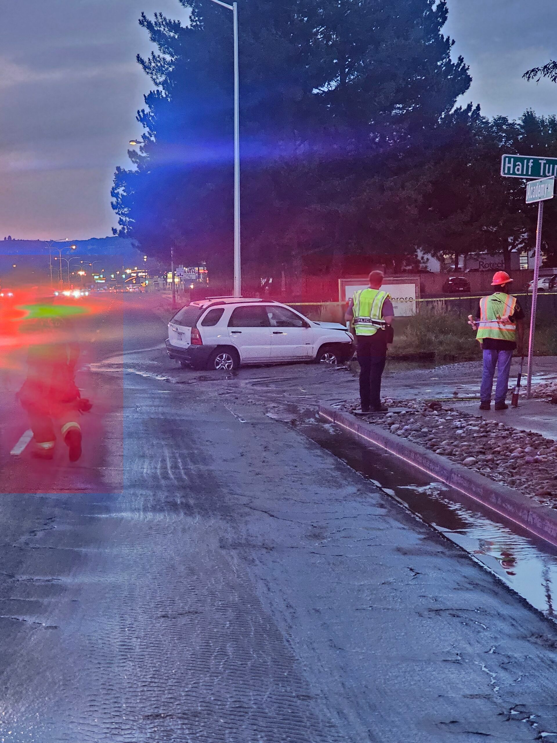 White SUV stuck in mud near a road, with two people in vests standing nearby, emergency lights visible.