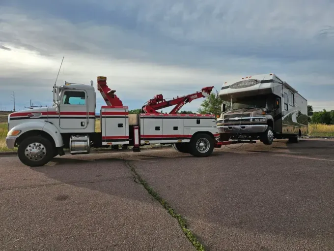 Tow truck towing a damaged RV on a paved road under a cloudy sky.
