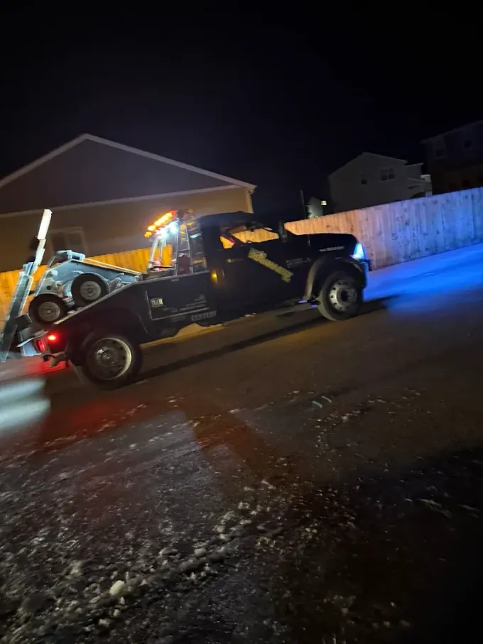 Tow truck at night, towing a car on a snowy, inclined road. Yellow lights on, dark background.