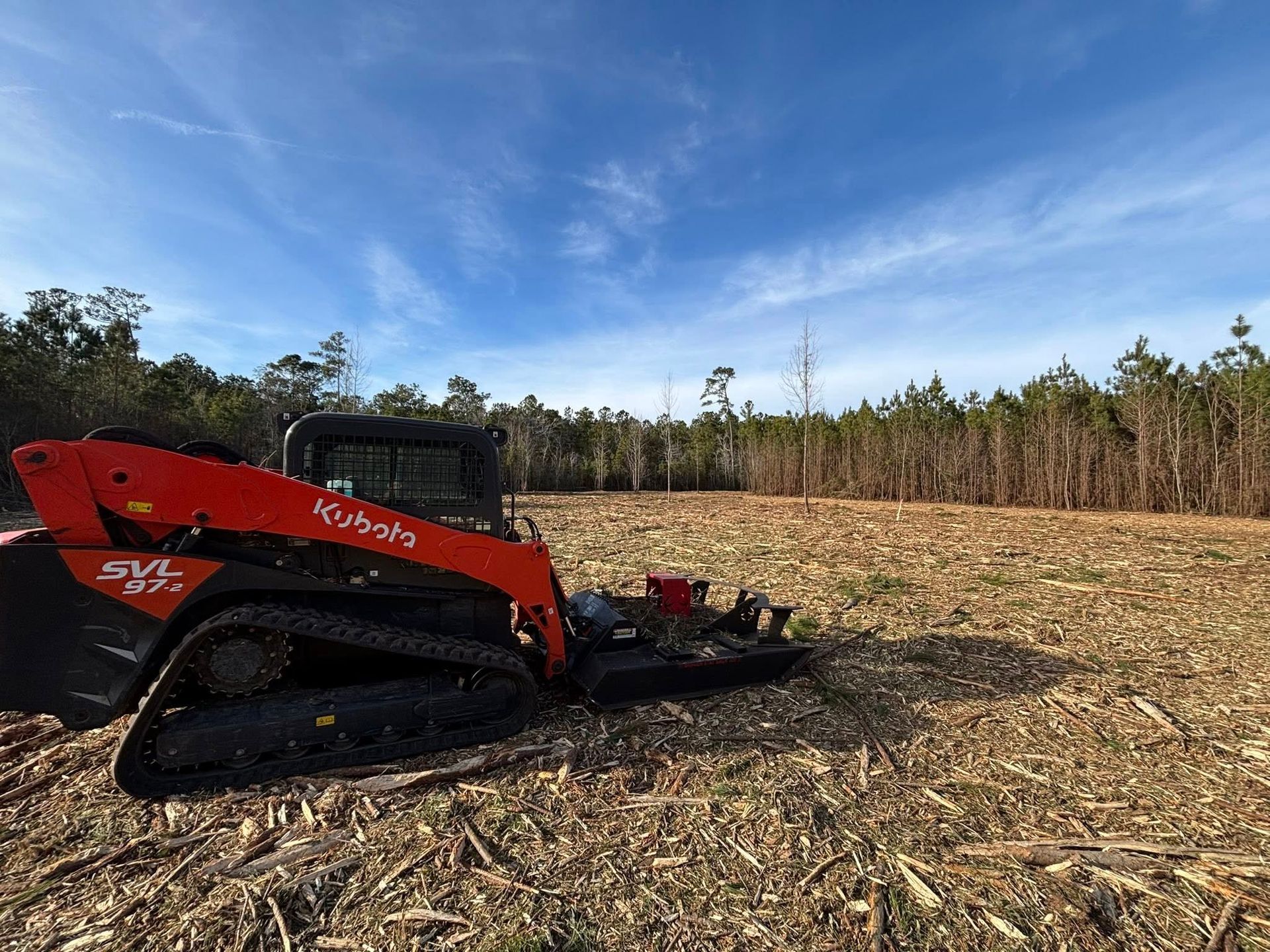 An orange Kubota SVL97-2 track loader with a brush cutter attachment sits on a recently cleared, wood-chipped forest lot.