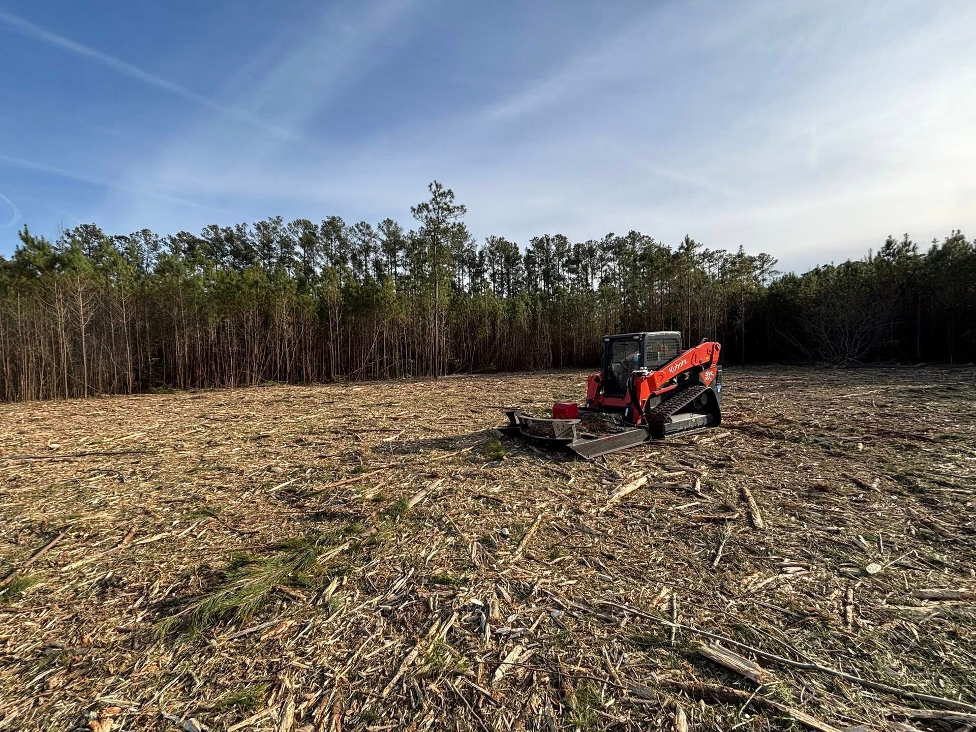 An orange mulcher tractor on a recently cleared field with a forest backdrop under a blue sky.