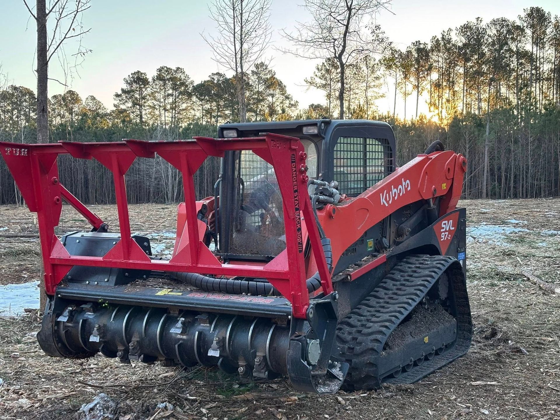 A red Kubota compact track loader equipped with a forestry mulcher attachment sits in a cleared wooded lot at sunset.